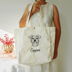 May include: Cream-colored tote bag with a black embroidered dog portrait and the name "Copper" in script. The bag has two side pockets and a snap closure. The tote is held by a person wearing a white dress.