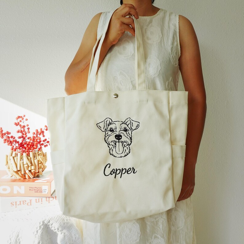 May include: Cream-colored tote bag with a black embroidered dog portrait and the name "Copper" in script. The bag has two side pockets and a snap closure. The tote is held by a person wearing a white dress.