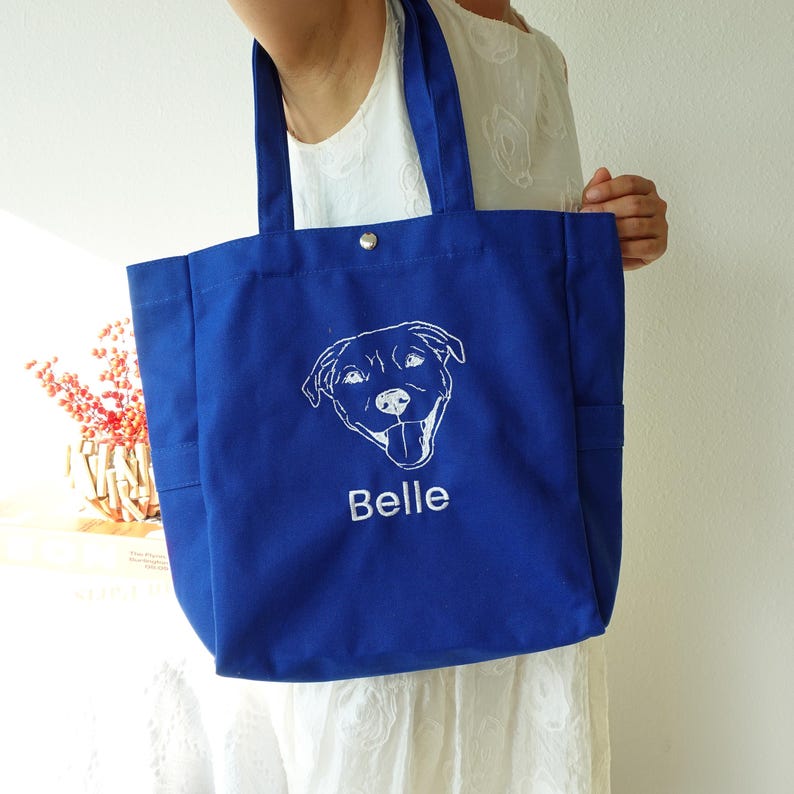 May include: A royal blue tote bag with a white embroidered dog face and the name "Belle" underneath. The bag has two side pockets and a snap closure at the top. The bag is made of a sturdy fabric.