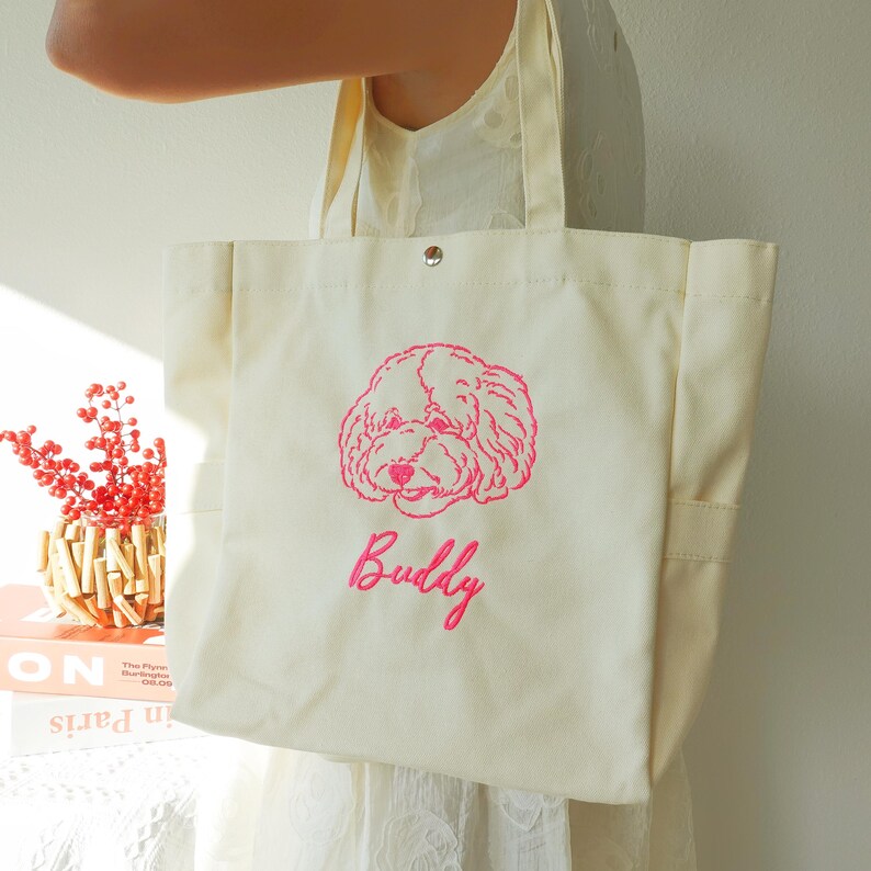 May include: Cream-colored tote bag with a pink embroidered illustration of a dog's face and the name "Buddy" in cursive. The bag has two handles and a button closure. The bag is made of a sturdy canvas material.
