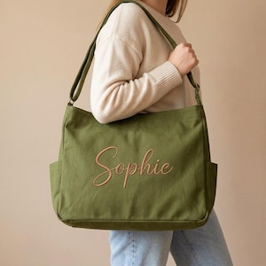 May include: Olive green canvas tote bag with the name "Sophie" embroidered in a cursive font. The bag has two exterior pockets and a shoulder strap. The person is wearing a cream-colored sweater and blue jeans.