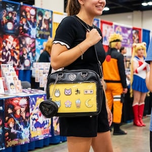 May include: Black crossbody bag with a yellow front panel featuring anime-themed pins. The bag has a small, heart-shaped coin purse attached. The background includes anime posters and cosplayers.