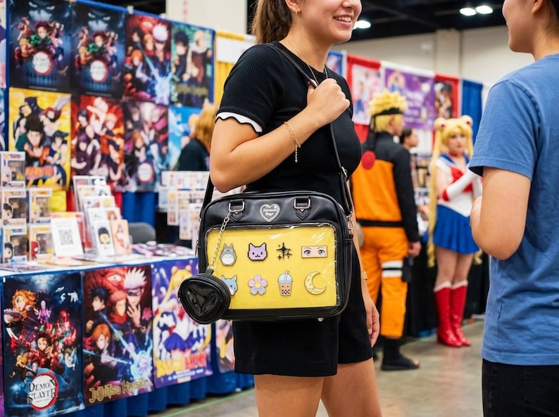 May include: Black crossbody bag with a yellow front panel featuring anime-themed pins. The bag has a small, heart-shaped coin purse attached. The background includes anime posters and cosplayers.