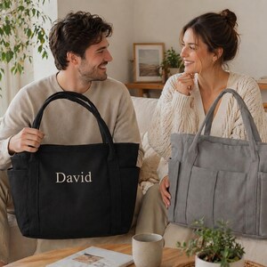 Puede incluir: Un bolso tote negro con el nombre "David" bordado en blanco. El bolso tiene dos asas y varios bolsillos exteriores. Tambi&eacute;n se ve un bolso tote gris con caracter&iacute;sticas similares.