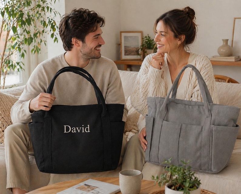 Puede incluir: Un bolso tote negro con el nombre "David" bordado en blanco. El bolso tiene dos asas y varios bolsillos exteriores. Tambi&eacute;n se ve un bolso tote gris con caracter&iacute;sticas similares.