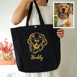May include: Black tote bag with a golden retriever design and the name "Buddy" in gold script. The bag has two side pockets and a snap closure. A small image of a golden retriever wearing a colorful bandana is in the upper right corner.