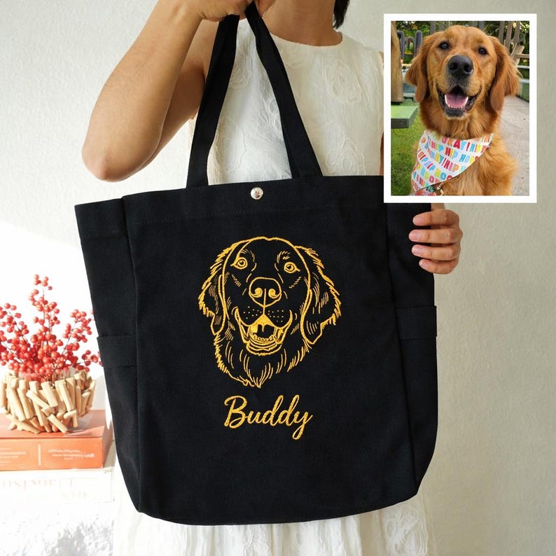 May include: Black tote bag with a golden retriever design and the name "Buddy" in gold script. The bag has two side pockets and a snap closure. A small image of a golden retriever wearing a colorful bandana is in the upper right corner.