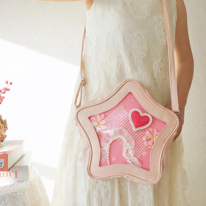 May include: A light pink star-shaped crossbody bag with a clear window. The window displays a pink and white polka dot pattern, a red heart, and floral accents. The bag has a long strap.