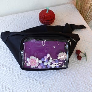 May include: Black fanny pack with a clear plastic window. The window shows a purple fabric with white and pink flowers and purple and white pom-poms.