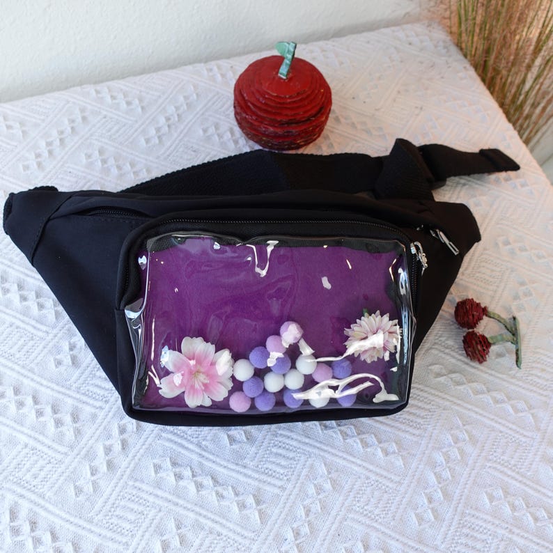 May include: Black fanny pack with a clear plastic window. The window shows a purple fabric with white and pink flowers and purple and white pom-poms.