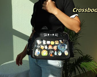 May include: Black crossbody bag with cat ears and a clear front panel filled with colorful pins. The bag has a white cat face design and a shoulder strap. The word "Crossbody" is in yellow text.