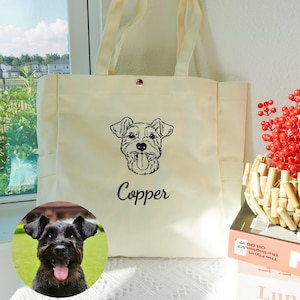 May include: Cream-colored tote bag with a black embroidered dog portrait and the name "Copper" below it. The bag has two handles and a snap closure. A small, round photo of a black dog is in the lower left corner.