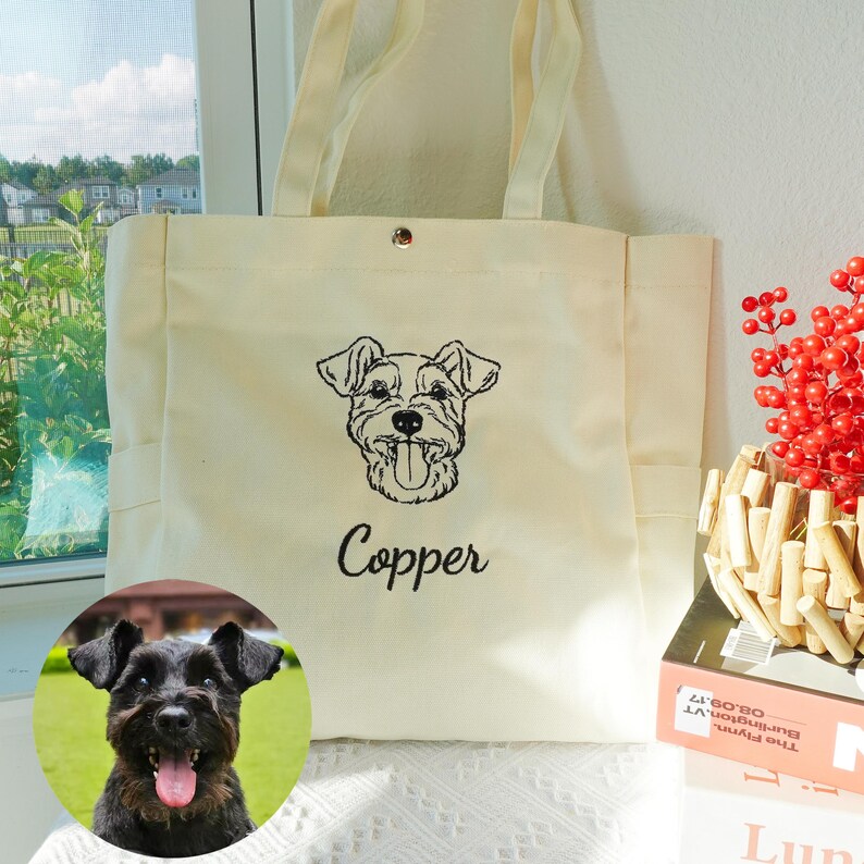 May include: Cream-colored tote bag with a black embroidered dog portrait and the name "Copper" below it. The bag has two handles and a snap closure. A small, round photo of a black dog is in the lower left corner.