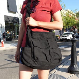 May include: A black canvas messenger bag with multiple front pockets is carried over the shoulder. The bag has a large main compartment and a flap closure. The strap is black. The person is wearing a red shirt and denim shorts.