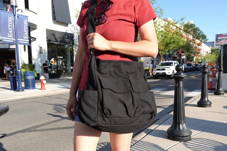 May include: A black canvas messenger bag with multiple front pockets is carried over the shoulder. The bag has a large main compartment and a flap closure. The strap is black. The person is wearing a red shirt and denim shorts.