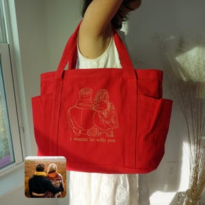 Puede incluir: Un bolso tote rojo con dos bolsillos exteriores y asas a juego. El bolso presenta una ilustraci&oacute;n dorada de un par de personas y el texto "I wanna be with you". La imagen en la esquina muestra una pareja abraz&aacute;ndose.