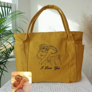 Puede incluir: Bolso tote mostaza con dos bolsillos exteriores y asas resistentes. El bolso presenta un dibujo lineal negro de una pareja abraz&aacute;ndose, con las palabras "I Love You" debajo. Una peque&ntilde;a foto de una pareja bes&aacute;ndose est&aacute; en la esquina inferior izquierda.