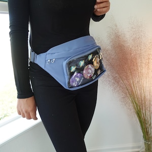 May include: A light blue fanny pack with a clear plastic window that shows a variety of colorful pins and patches. The fanny pack is worn around the waist of a person.