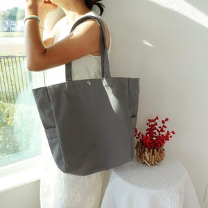 May include: A gray canvas tote bag with two exterior pockets and a snap closure. The bag has two long handles for carrying over the shoulder. The tote is being held by a person wearing a white dress.