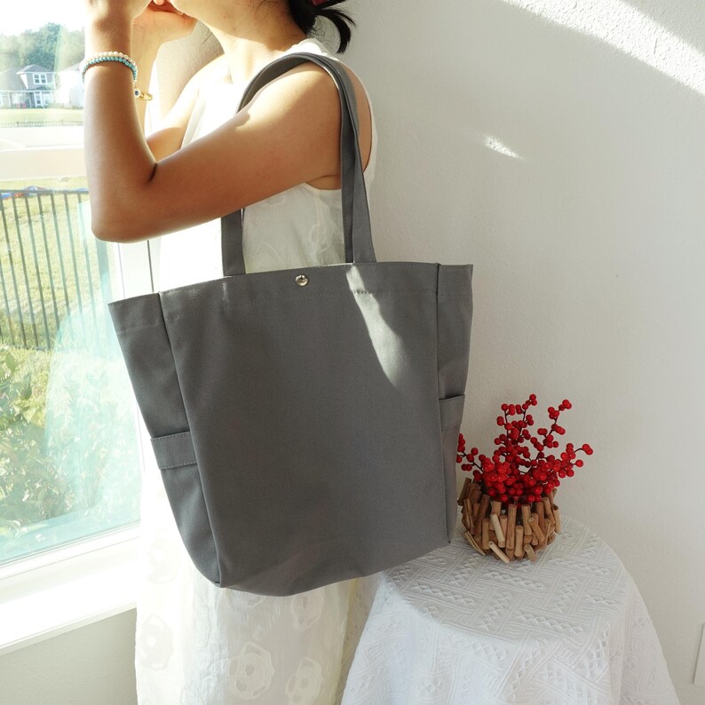 May include: A gray canvas tote bag with two exterior pockets and a snap closure. The bag has two long handles for carrying over the shoulder. The tote is being held by a person wearing a white dress.