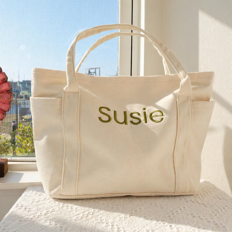 Puede incluir: Bolso tote de lona color crema con dos bolsillos exteriores y dos asas. La palabra "Susie" est&aacute; bordada en verde en la parte delantera de la bolsa. La bolsa est&aacute; sobre un mantel de encaje blanco.