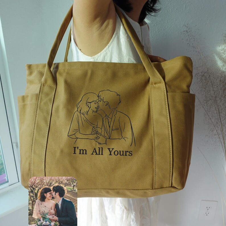 Puede incluir: Un bolso tote de lona color canela con dos bolsillos exteriores y asas largas. La bolsa presenta un dibujo lineal negro de una pareja bes&aacute;ndose, con el texto "I'm All Yours" debajo. Una peque&ntilde;a foto de la pareja est&aacute; en la esquina inferior izquierda.