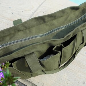 Puede incluir: Bolso tote de lona verde oliva con cremallera superior y m&uacute;ltiples bolsillos exteriores. El bolso tiene asas resistentes y un forro de color neutro. El bolso est&aacute; sobre una superficie clara.