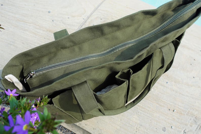 Puede incluir: Bolso tote de lona verde oliva con cremallera superior y m&uacute;ltiples bolsillos exteriores. El bolso tiene asas resistentes y un forro de color neutro. El bolso est&aacute; sobre una superficie clara.