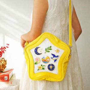 May include: A yellow star-shaped shoulder bag with a white interior and decorative pins. The pins include a crescent moon, a blue bird, a flower, a compass, and other nature-themed designs. The bag has a long yellow strap.