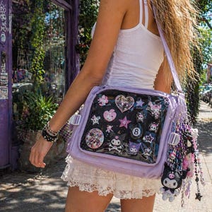 Puede incluir: Un bolso mensajero lila con un panel frontal transparente que muestra varios amuletos decorativos. El bolso tiene una correa larga y est&aacute; adornado con un amuleto de cuentas. El bolso se lleva al hombro.