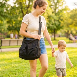 May include: A black shoulder bag with multiple front pockets is carried by a person walking in a park with a child. The bag has a wide shoulder strap. The scene is outdoors on a sunny day.