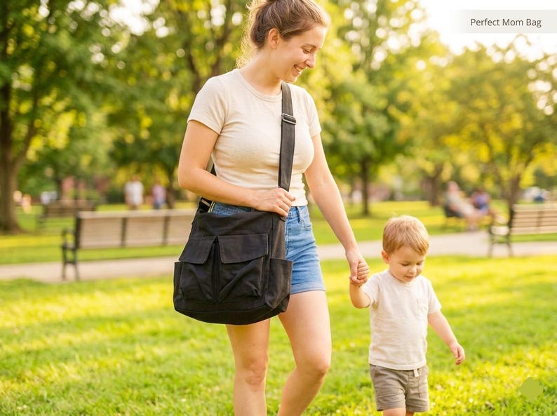 May include: A black shoulder bag with multiple front pockets is carried by a person walking in a park with a child. The bag has a wide shoulder strap. The scene is outdoors on a sunny day.