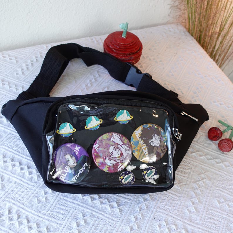 May include: Black fanny pack with a clear plastic front panel. The panel has several round pins with anime characters and designs. The pins include a blue and white pin with the text "BLUE" and "VIL-D-77", a pink and purple pin with a female character, a black and white pin with a male character, and two green and white pins with a planet design.