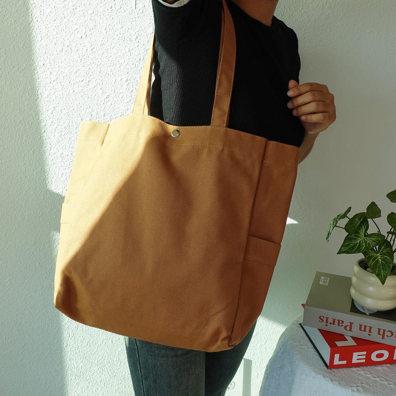 May include: A tan canvas tote bag with two handles and a snap closure. The bag has a rectangular shape with a pocket on the front. The person is wearing a black shirt and blue jeans.