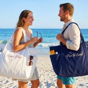 Puede incluir: Dos personas en una playa sostienen cada una una bolsa de mano grande. Una bolsa es blanca con "Beach Day" bordado en rosa. La otra es azul marino con "Pun & Sand" bordado en blanco. Ambas bolsas tienen bolsillos.