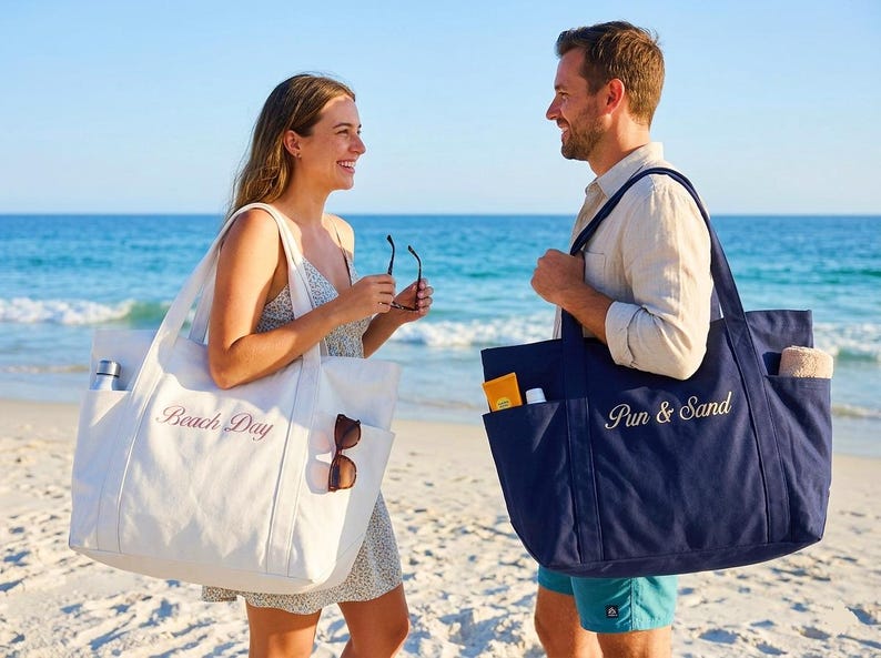 Puede incluir: Dos personas en una playa sostienen cada una una bolsa de mano grande. Una bolsa es blanca con "Beach Day" bordado en rosa. La otra es azul marino con "Pun & Sand" bordado en blanco. Ambas bolsas tienen bolsillos.