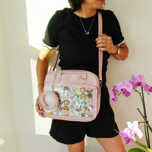 May include: A light pink messenger bag with a clear front pocket filled with colorful pins. The bag has a matching small coin purse attached. The bag is being held by a person.