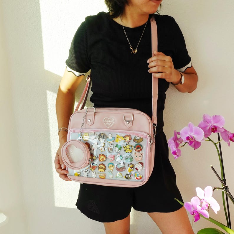May include: A light pink messenger bag with a clear front pocket filled with colorful pins. The bag has a matching small coin purse attached. The bag is being held by a person.