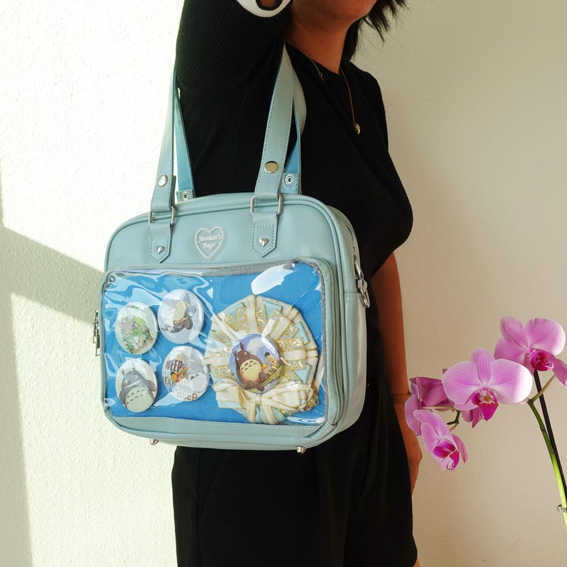May include: A light blue satchel bag with a clear front pocket displaying several round pins and a decorative ribbon. The bag has a heart-shaped emblem and silver hardware. The bag is carried by a person wearing a black top.