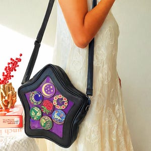 May include: Black star-shaped crossbody bag with a clear front panel displaying colorful circular pins. The pins feature various designs, including moons, flowers, and birds, set against a purple background. The bag has a black strap and trim.
