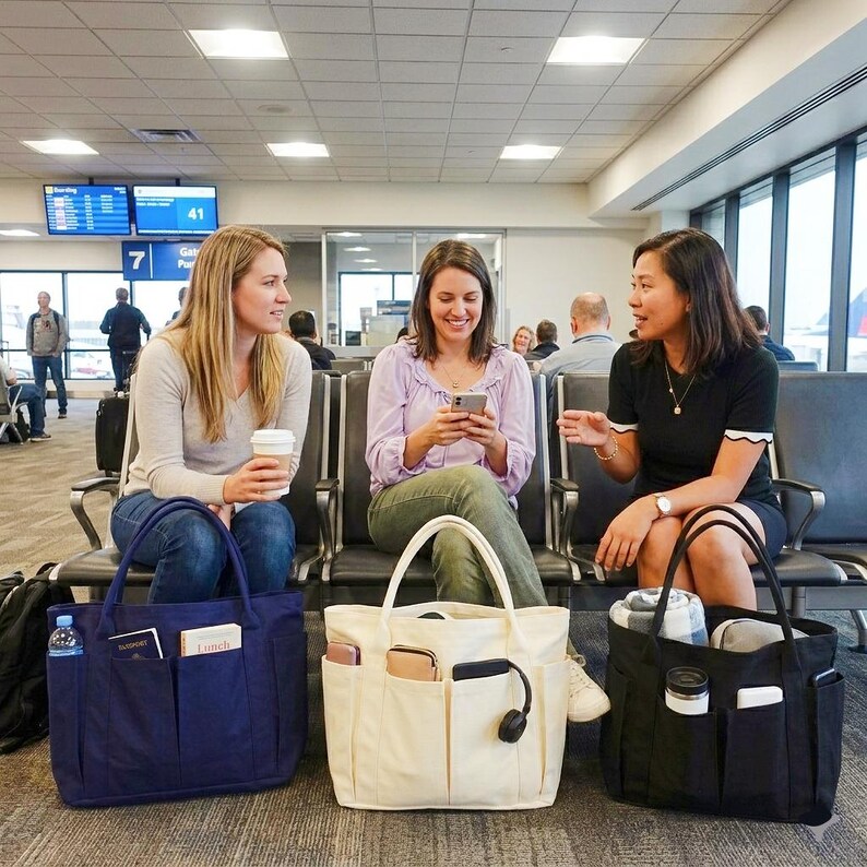 Puede incluir: Tres mujeres en un aeropuerto, cada una con un bolso de mano. Un bolso de mano azul marino, un bolso de mano color crema y un bolso de mano negro. Los bolsos tienen m&uacute;ltiples bolsillos y est&aacute;n llenos de art&iacute;culos como botellas de agua, auriculares y pasaportes.