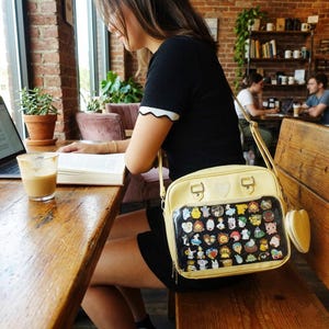 May include: A pale yellow crossbody bag with a clear front panel displaying various character pins. The bag has a matching heart-shaped coin purse attached. The person is sitting at a wooden table in a cafe.