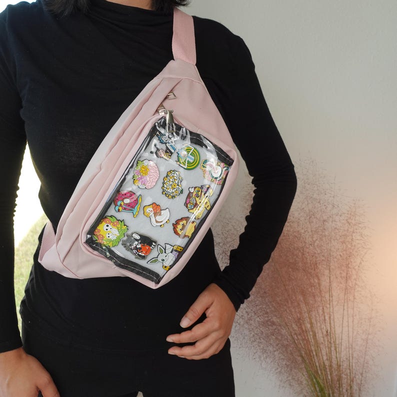 May include: A pink fanny pack with a clear plastic window on the front. The window shows a variety of colorful enamel pins.