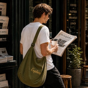 May include: A person wearing a white t-shirt and black pants, carrying a green shoulder bag with the word "Explore" embroidered on it. The person is holding a newspaper and a coffee cup near a newsstand with newspapers.