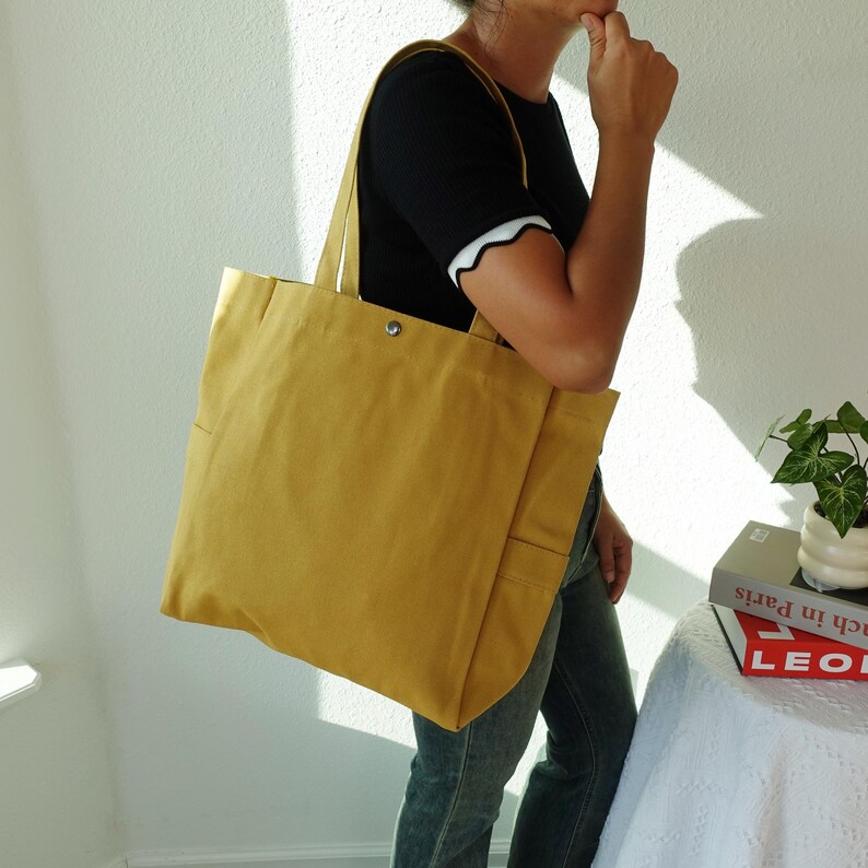 May include: Mustard yellow tote bag with shoulder straps. The bag has a rectangular shape with a snap closure and a pocket on the front. The bag is carried over the shoulder.