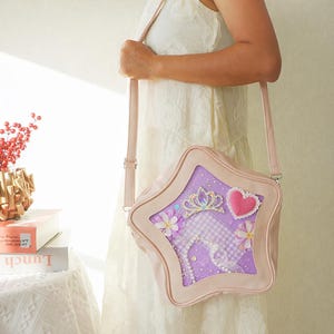 May include: A light pink star-shaped crossbody bag with a clear window displaying a purple interior. The interior features a crown, heart, flowers, and a checkered pattern. The bag has a long, adjustable strap.