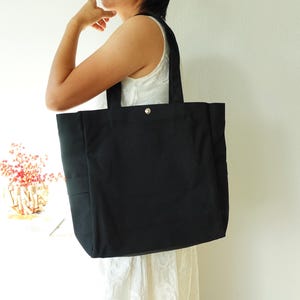 May include: A large, black tote bag with sturdy handles and a silver snap closure. The bag is made of a durable fabric and features a simple, minimalist design, suitable for carrying various items. The bag has a rectangular shape and a side pocket.