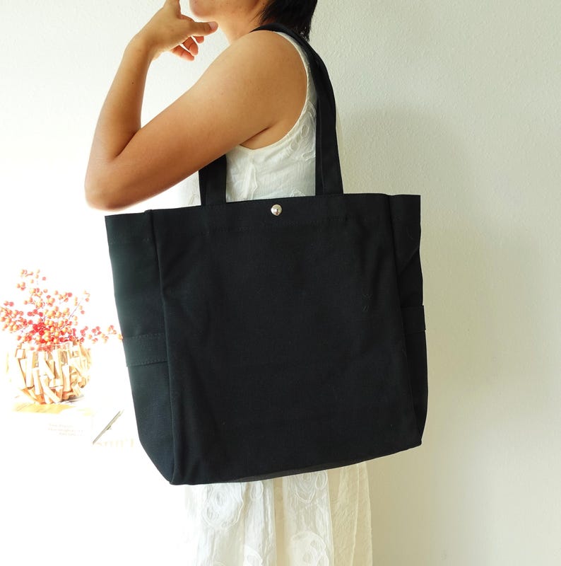 May include: A large, black tote bag with sturdy handles and a silver snap closure. The bag is made of a durable fabric and features a simple, minimalist design, suitable for carrying various items. The bag has a rectangular shape and a side pocket.