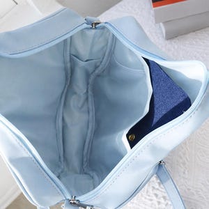 May include: Open view of a light blue handbag with a zipper closure. The interior reveals a light blue lining and several pockets. A dark blue item is partially visible inside the bag.