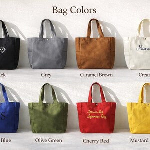 May include: A collection of eight tote bags in various colors, including black, grey, caramel brown, cream, royal blue, olive green, cherry red, and mustard yellow. Each bag has a rectangular shape with two handles and a pocket. Some bags have embroidered text.